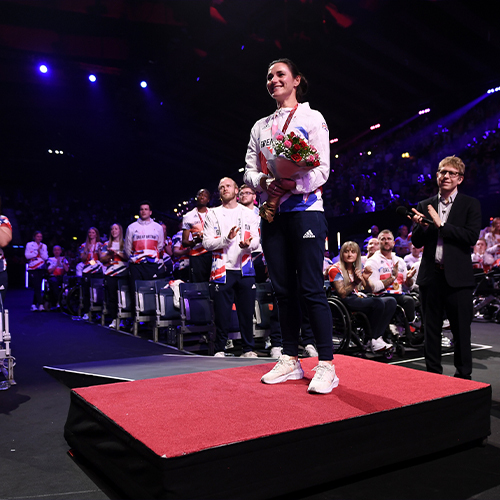 A woman proudly stands on a stage, holding a trophy, at the Paralympics Team GB's national lottery homecoming event at AO arena.